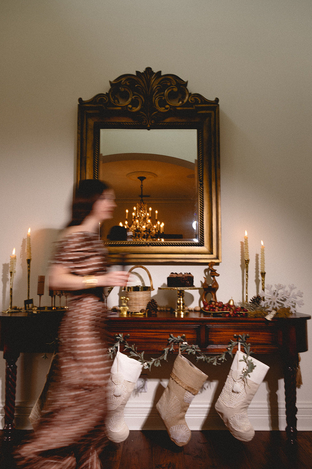 Woman in a patterned dress standing in a room with stockings on a mantel and a large mirror reflecting a chandelier.